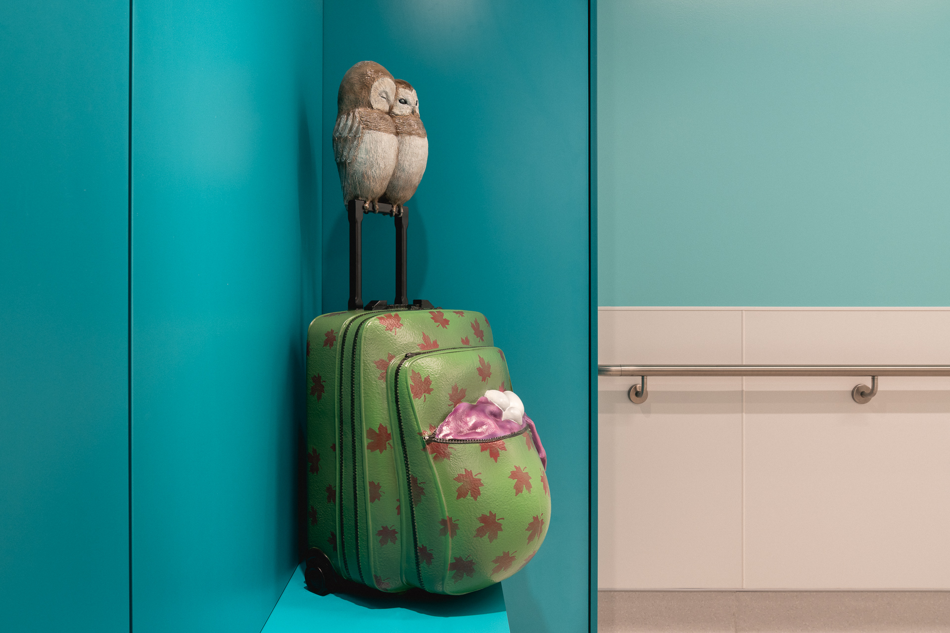 Deux chouettes, appuyées l’une sur l’autre, sont posées sur la poignée d’une valise à l’accueil de la maternité. Dépassant d’une poche de la valise, trois œufs sont nichés dans une manche d’un chandail. Bien remplie, la valise a la forme arrondie du ventre d’une femme enceinte.